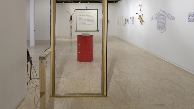 Art gallery installation featuring a red barrel, a framed sign labeled "ARTS MEDIA SPORTS," and various wall-mounted mixed-media pieces, including text-covered fabric and sculptural elements.