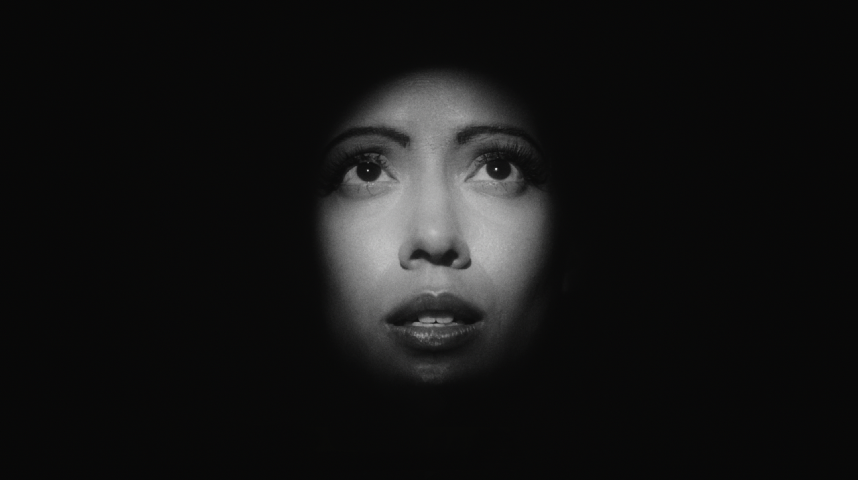 Black-and-white close-up portrait of a face emerging from darkness. Only the eyes, nose, and slightly parted lips are illuminated by a spotlight while the edges of the face fade into shadow. The upward gaze conveys vulnerability or apprehension, combing Hollywood glamour lighting with a sense of psychological distress.