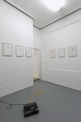 tape recorder on a grey floor with cables extending out from it. on the walls is a series of framed works.