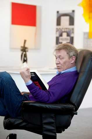 man sat in a leather office chair reading Finnegan's Wake by James Joyce. Behind him there are canvases on the wall.