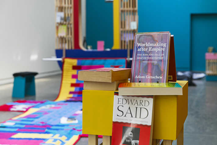 A brighly lit room with a shelving unit in the foreground displaying a number of books. In the background is a brightly coloured tapestry on the floor.