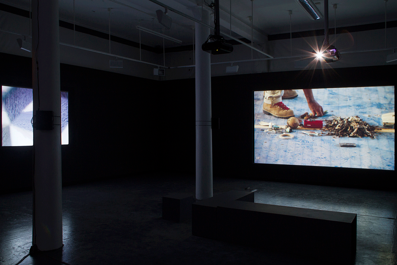 In a dark room, there are two projector screens on walls perpendicular to each other. The screen on the right shows a pair of shoes and a hand picking up an object from the ground. There are multiple objects on the ground, including a heap of leaves, a ball, tape and a cassette tape. There are two pillars placed in through the centre of the room. A bench is placed next to the pillar near the screen on the right. The screen on the left side has an image of a water body in different lighting.