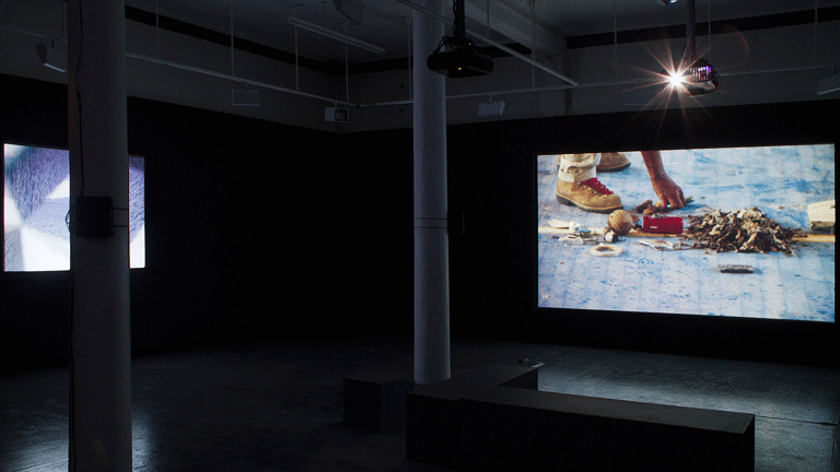 In a dark room, there are two projector screens on walls perpendicular to each other. The screen on the right shows a pair of shoes and a hand picking up an object from the ground. There are multiple objects on the ground, including a heap of leaves, a ball, tape and a cassette tape. There are two pillars placed in through the centre of the room. A bench is placed next to the pillar near the screen on the right. The screen on the left side has an image of a water body in different lighting.