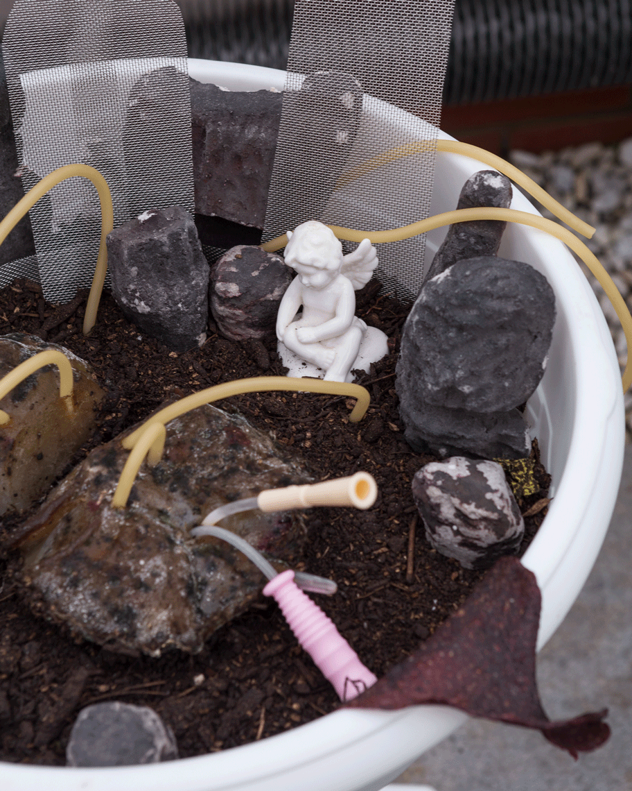 Close up view of a small sculptural figure of an angel in a plant pot with tubes coming in and out of the soil 