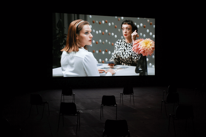 An image of a film being screened in a dark room. The screen shows two people sat at a table, one appears to be giving the other a tarot reading. They both look surprised. There are eggs suspended in the air behind them and dahlias in a vase on the table.