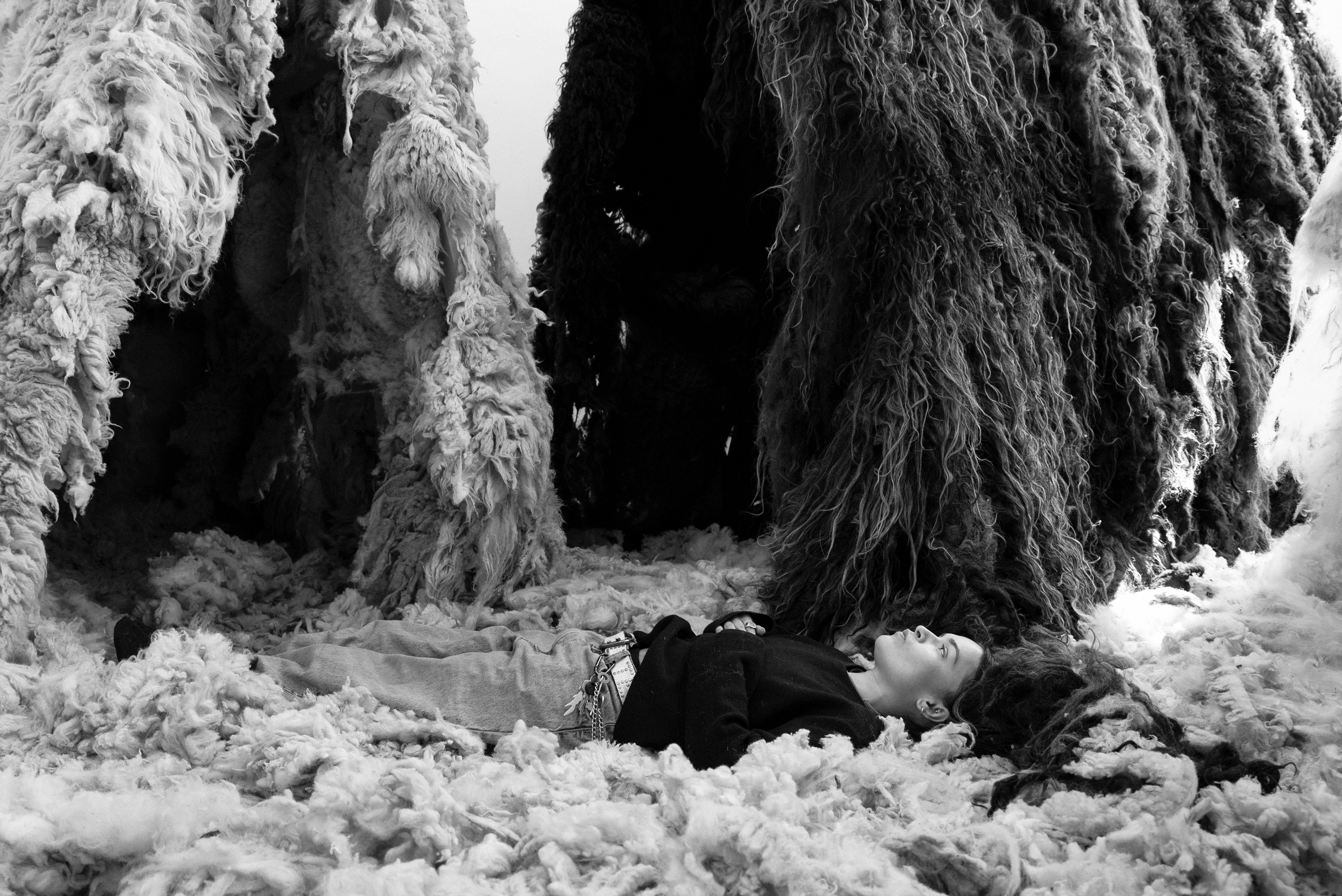  A black-and-white photograph of the artist Oliver Priya Foster lying on thick sheep’s wool with wool covered structures in the background. 