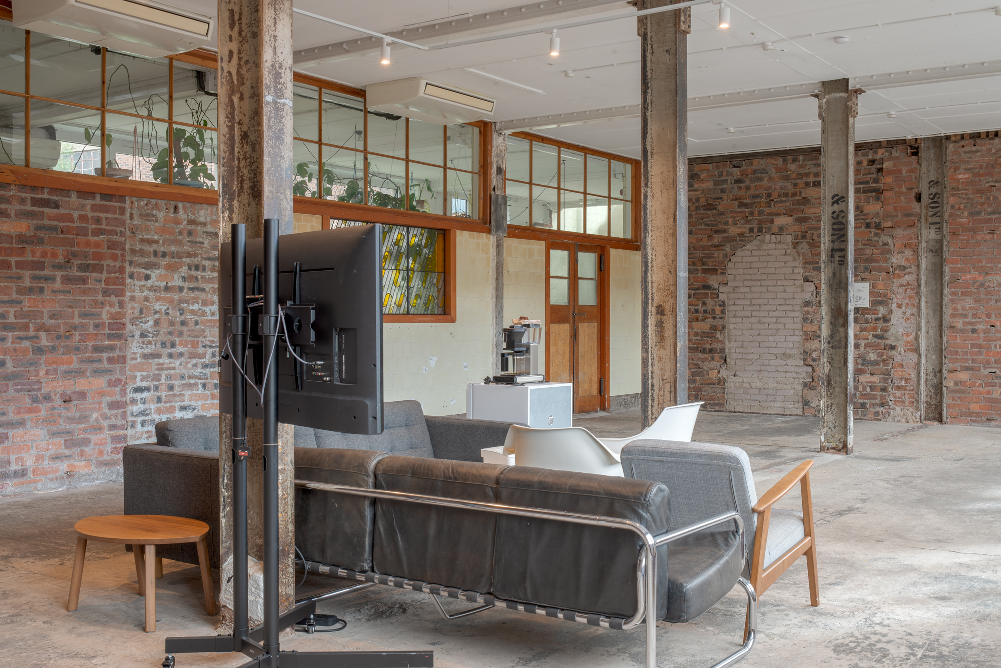 View through an industrial space with steel columns and mixed brick and tiled walls, showing a seating area with sofas and a wall-mounted television, a stained-glass window and wooden door visible beyond.