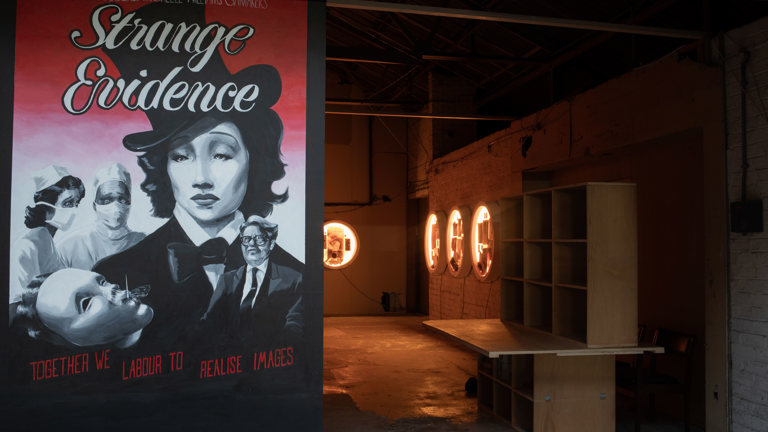A painted vintage-style film poster reading "Offline presents Michelle Williams Gamaker's Strange Evidence, together we labour to realise images," beside an entrance into a dim warehouse gallery lit by circular illuminated display cases.