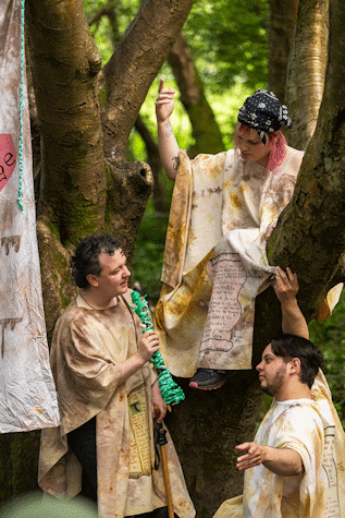 Three people wearing off white cloaks stand near, and sit on, a tree. One is holding a wooden rod wrapped in rope.
