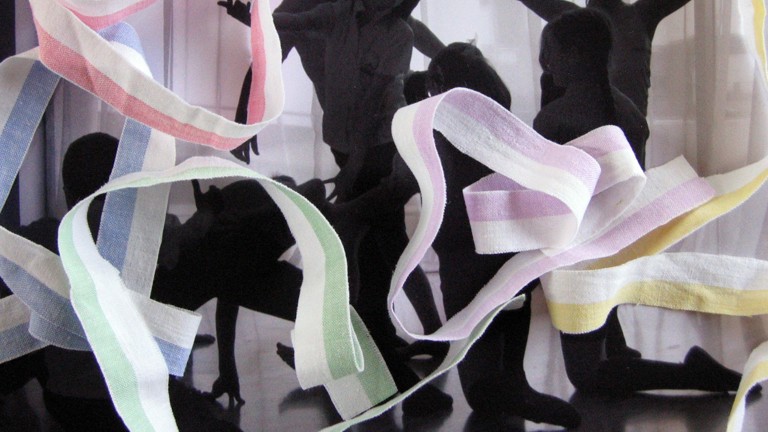 A black and white photograph has been printed and then scanned again, with colourful ribbons of fabric arranged over the top, obscuring parts of the photo. In the photo a group of children are silhouetted against a curtained window, striking poses and dancing.