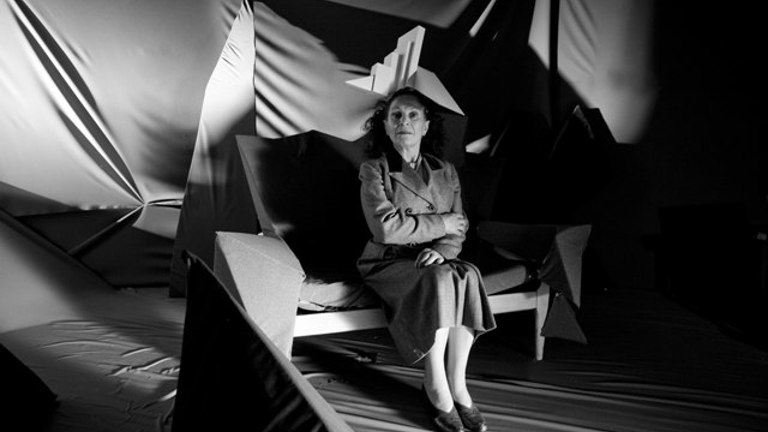 black and white film still of a woman sat on a bench with fabric hanging sculpturally behind