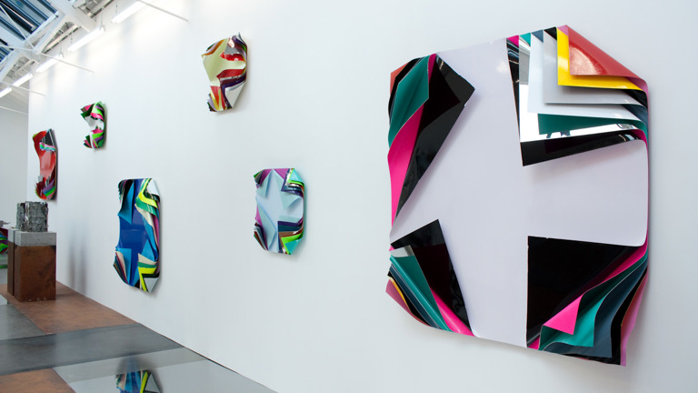 Installation view of colorful, abstract wall sculptures with layered, folded surfaces on a white gallery wall.