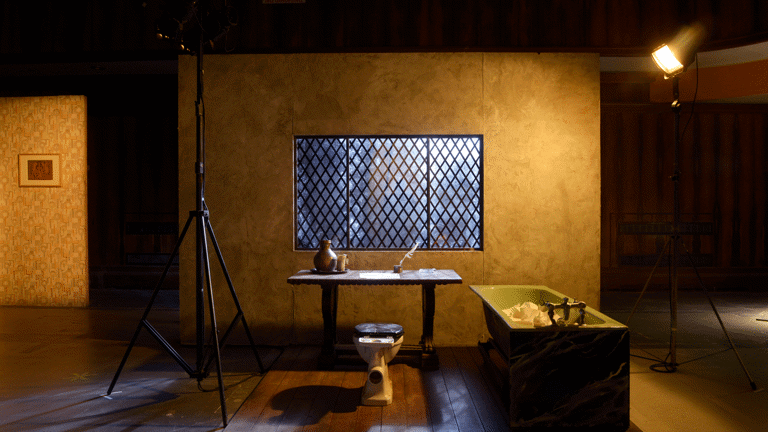 A wooden board floor has on top a wooden desk and a toilet as a chair with a green bathtub to the right. There is a wall with a window with diamond-shaped metal bars. It is all illuminated by two big spotlights with a warm yellow light. There are other sets behind and around this one. This is part of an art installation in Film City, Glasgow International 2018. 