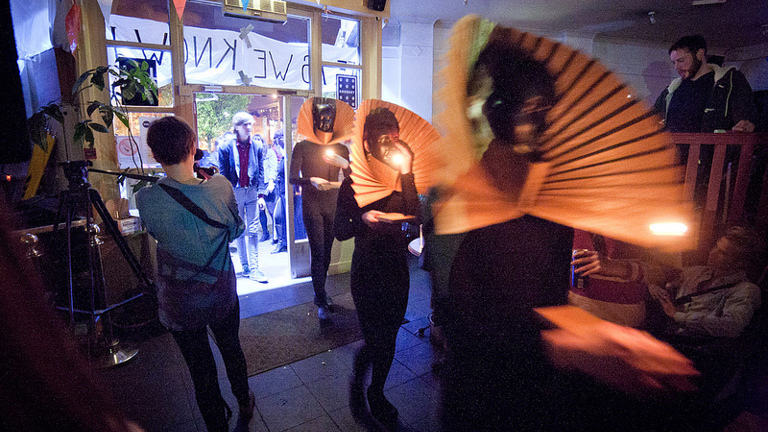 three performers in all black with large pleated collars holding torches enter a room from outside as onlookers watch.