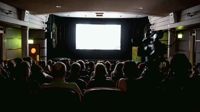 In a dark theatre, there is a projector screen with a blank screen at the front. There are theatre chairs placed opposite the screen, all the seats are full with people apart from the one at the centre of the back row. There are doors on the two side walls which are perpendicular to the screen. There is a video camera placed behind the back row of the theatre seats.