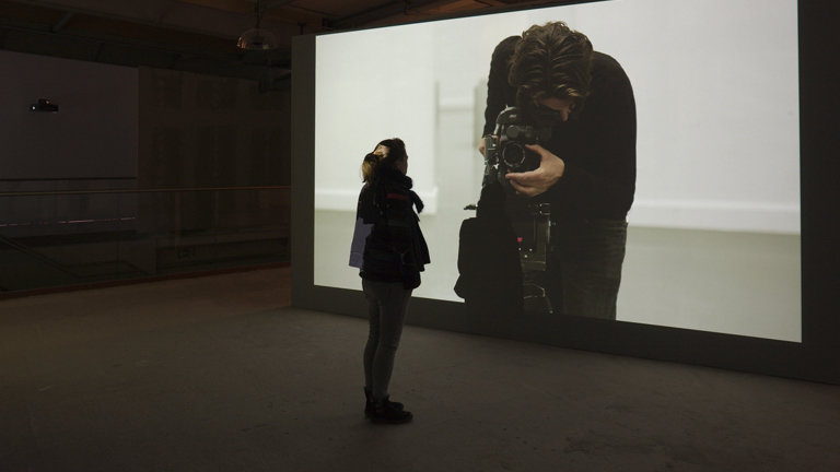 a person is stood in front of a large projection screen. on the screen there is a person looking into a viewfinder taking a photograph 