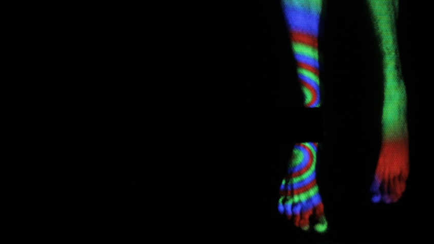 In a dark background, some legs are dangling and the holographic colorful lights are reflected onto them.