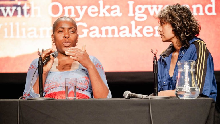 Close-up of two women in conversation on a panel stage, one speaking into a microphone with her hand raised, seated beside a table with water glasses and a jug.