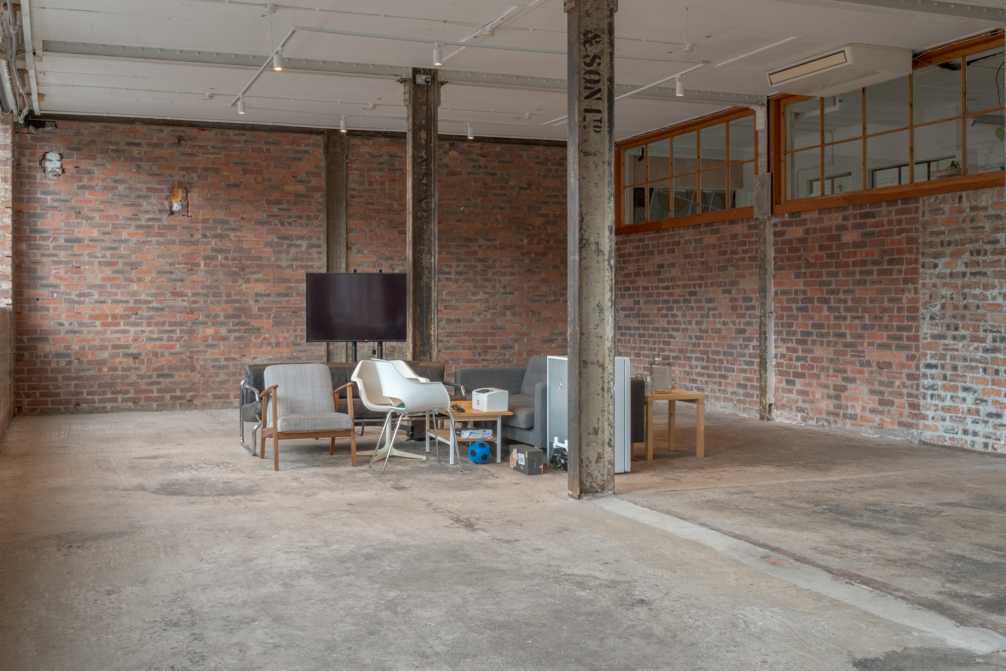 Wide view of an empty industrial space with exposed brick walls and steel columns, a small furnished seating area with sofa, chairs and television positioned in one corner beneath a mezzanine window.