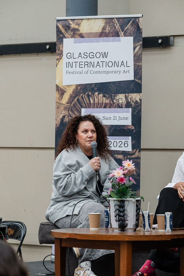 A woman with curly hair, wearing a grey tracksuit, sits speaking into a microphone during a panel discussion, with a Glasgow International festival banner behind her and a small table of flowers and drinks in front.