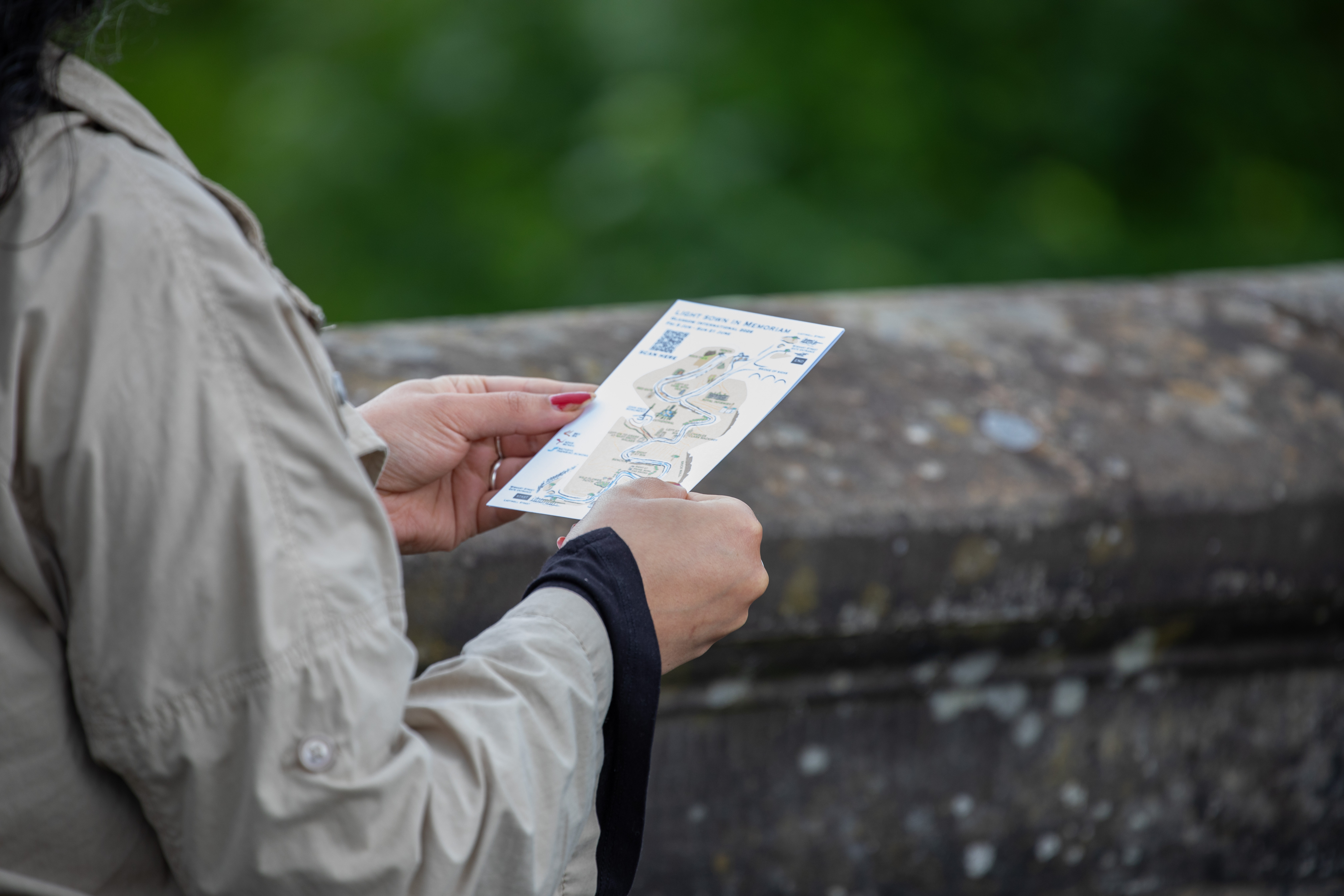 Close-up of a person's hands holding a printed trail map titled "Light Sown in Memoriam" against a mossy stone wall, greenery blurred in the background.