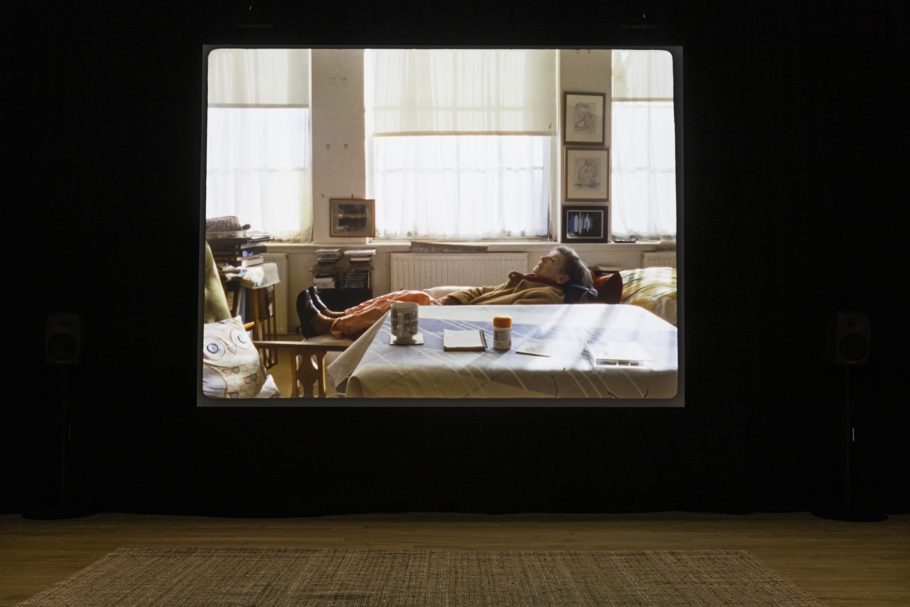 Darkened gallery with a large projected film still of an elderly person reclining on a sofa in a sunlit living room, surrounded by framed drawings, books, and a table with a mug and pill bottle.