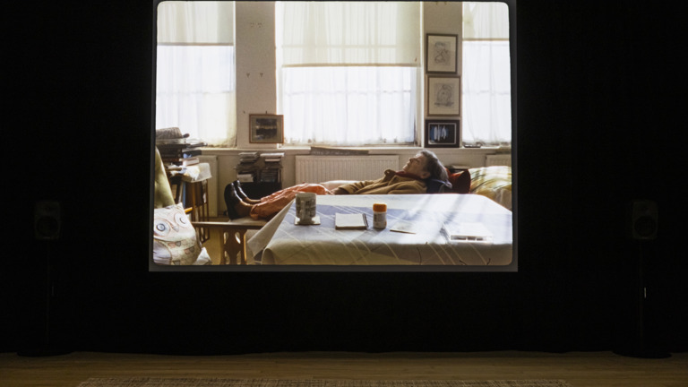Darkened gallery with a large projected film still of an elderly person reclining on a sofa in a sunlit living room, surrounded by framed drawings, books, and a table with a mug and pill bottle.