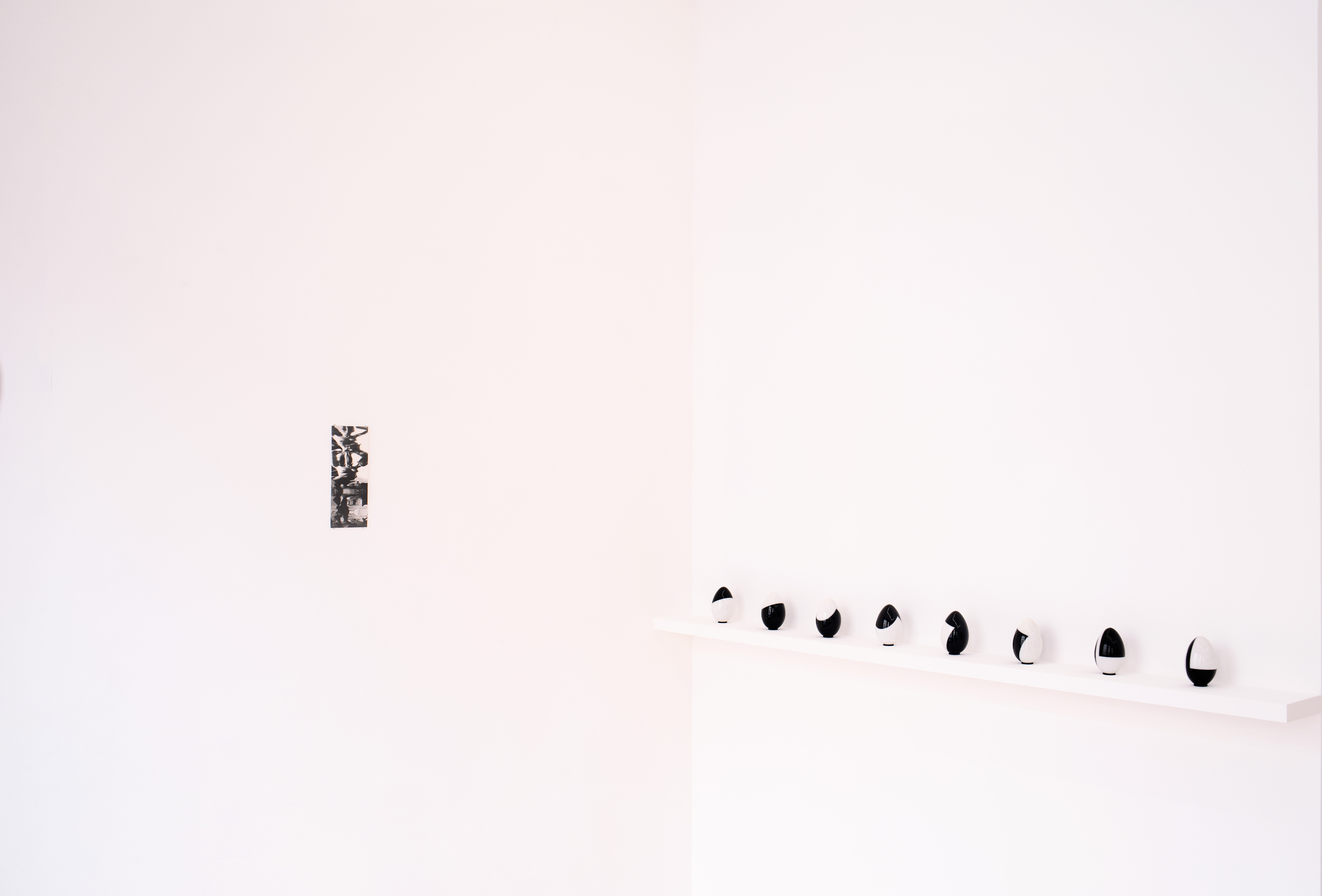 Minimalist gallery corner with a single small black-and-white photograph on the left wall and a row of black-and-white painted egg-shaped sculptures on a shelf along the adjoining wall.