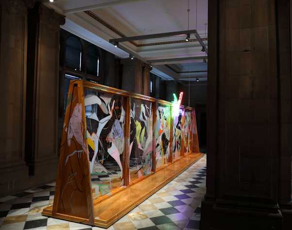 An abstract neon light showing two legs kicking the air in pointed heels, hangs at the top of a large wooden structure with glass panels and covered in fast gestural line paintings of nude figures and faces
