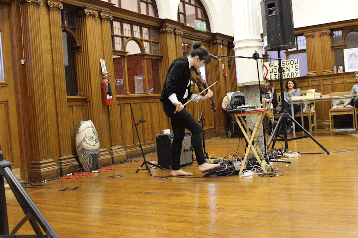 A woman is playing the violin, barefoot, in an old fashioned wooden room. There is musical equipment placed around her.