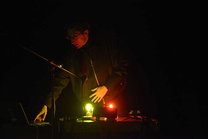 A dark photograph with a person dimly lit, they are stood at a desk which is lit by orange and green lights.