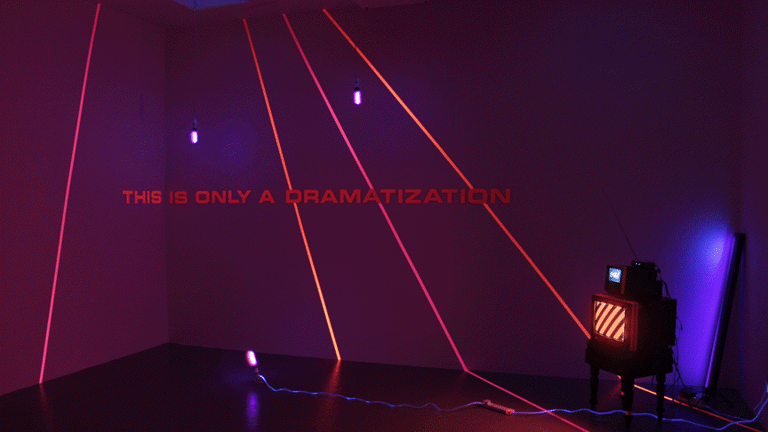 dark room with red and blue lighting. red laser like lines are projected across the wall along with he words 'THIS IS ONLY A DRAMATIZATION'. in the corner of the room is a small cube monitor with an even smaller monitor resting on top.