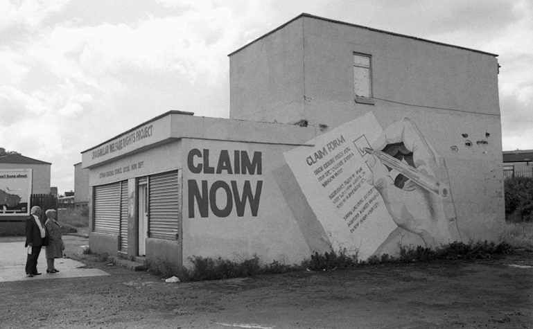 A black and white image of an outdoor area featuring a building. On the side of the building has been painted with the text 'claim now' and a hand signing a ballot paper. A main and a woman stand looking at the building.