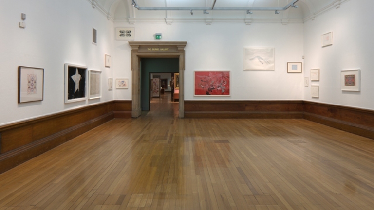 large room with wooden floors and white walls. installed on the walls at different heights are various framed works on paper.