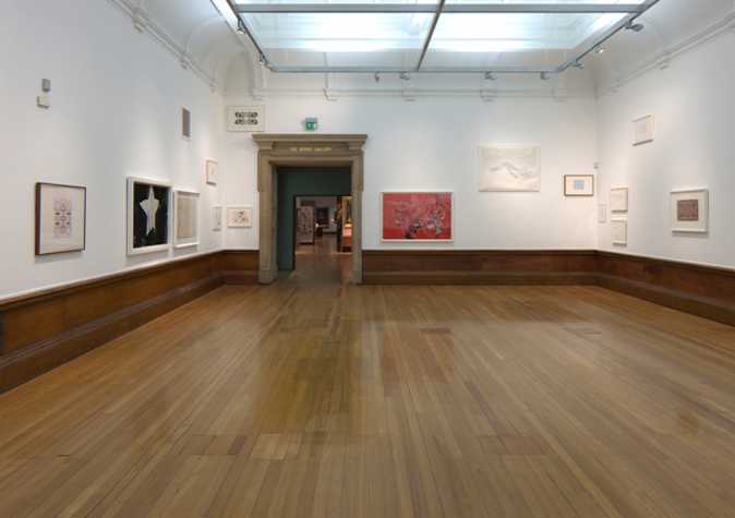 large room with wooden floors and white walls. installed on the walls at different heights are various framed works on paper.