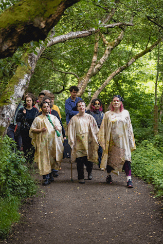 Three people wearing off white cloaks lead a group of people through a woodland.