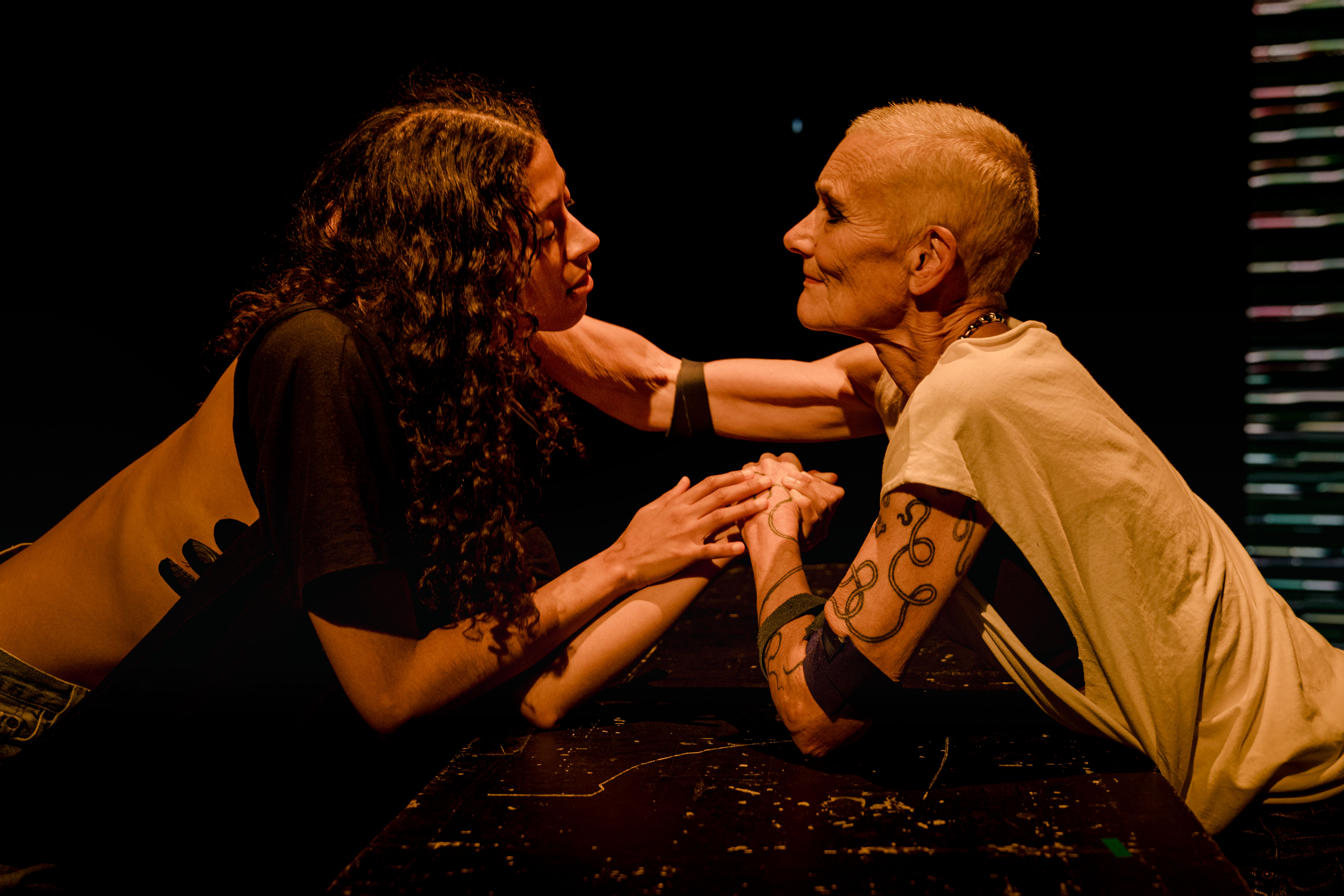 Two performers arm wrestle over a heavily marked black table in a black-box performance space. The performer on the left has long black curly hair, while the performer on the right has short blonde hair, a snake tattoo on their arm, and wears a white vest.