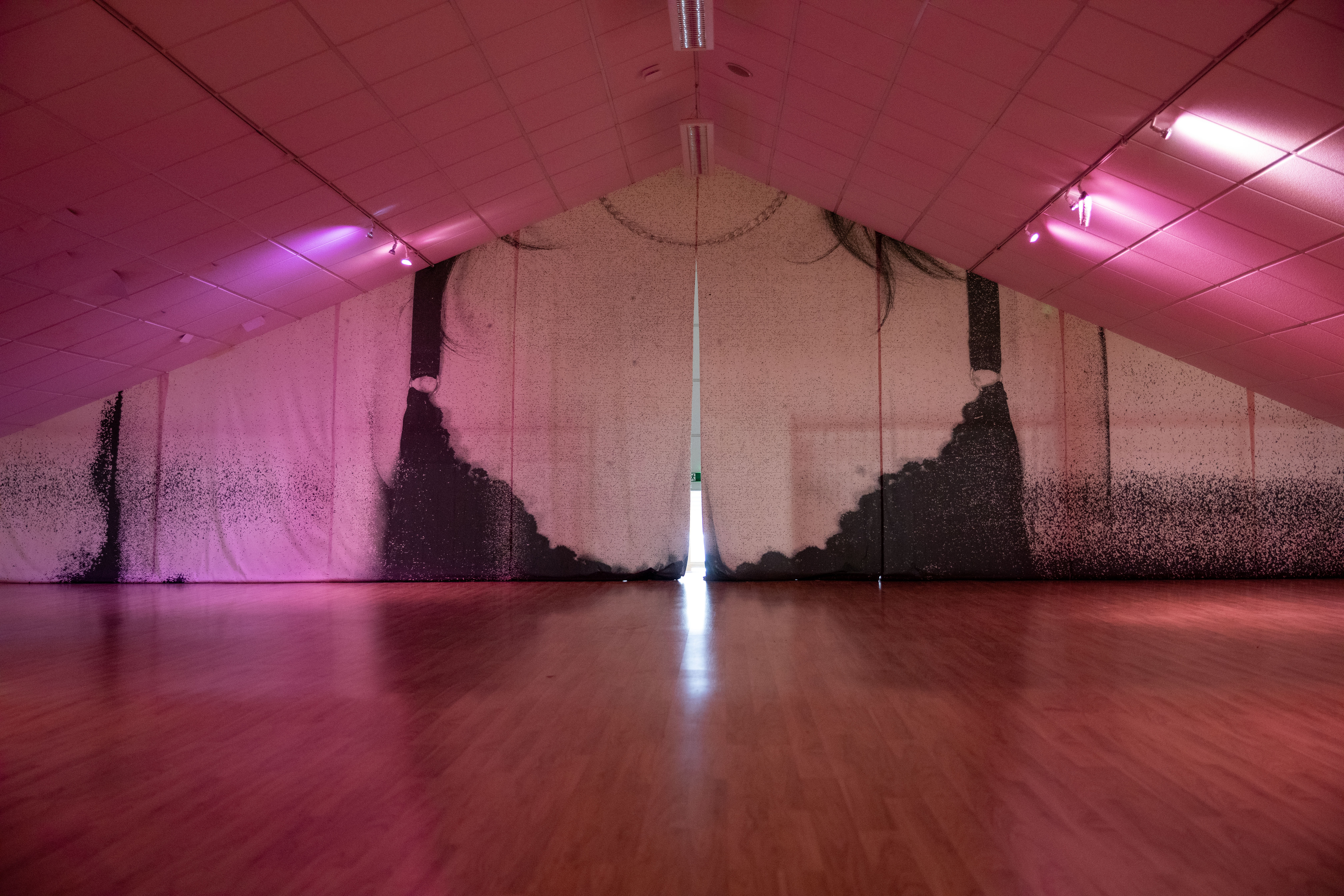 A large fabric backdrop hangs across an attic-style gallery at Gulabi Gallery, printed with a faded silhouette of hanging hair and a chain necklace, lit in pink, with a gap of light showing through the centre.