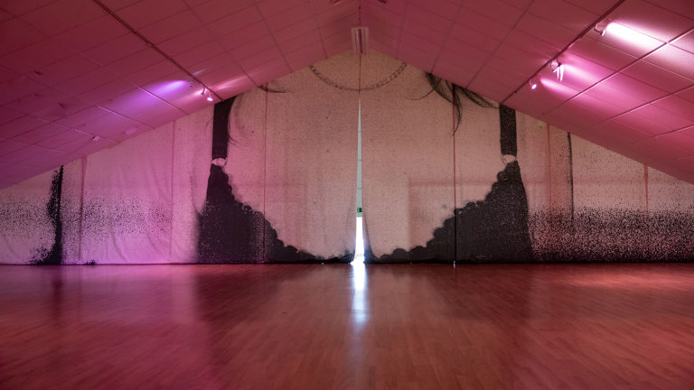 A large fabric backdrop hangs across an attic-style gallery at Gulabi Gallery, printed with a faded silhouette of hanging hair and a chain necklace, lit in pink, with a gap of light showing through the centre.