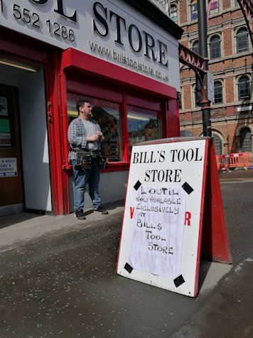 There is a sign outside Bill's Tool Store that reads "L'Outil Now Avalable Exclusively at Bill's Tool Store". There is a white man with a flannel and jeans with a tool belt in front of the store.