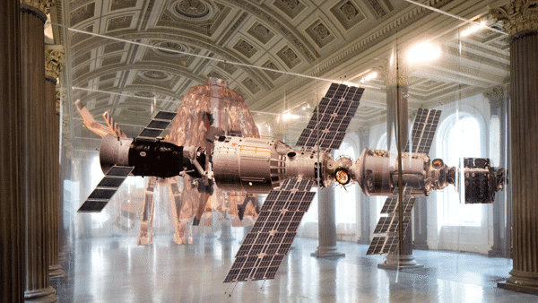 The image of a satellite can be seen printed on polyester foil panels, behind which there is another wall of polyester foil panels with a printed coat. It is hanging from a clothes hanger. Behind the coat, there is another wall of polyester foil panels on which you can see the printed legs of a robot. 