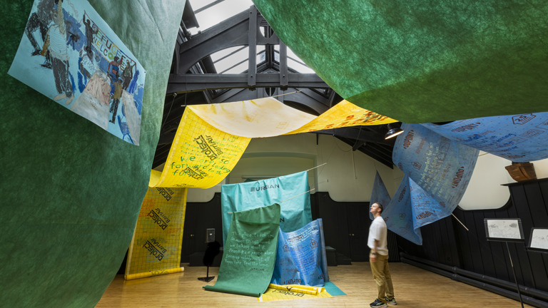 Installation view of an exhibition featuring large hanging fabric banners in green, yellow and blue printed with handwritten text and drawings, including a tent-like structure reading "EURBAN" and a painting of figures holding a "REFUGEE WELCOME" sign, with a visitor looking up at the hanging works.