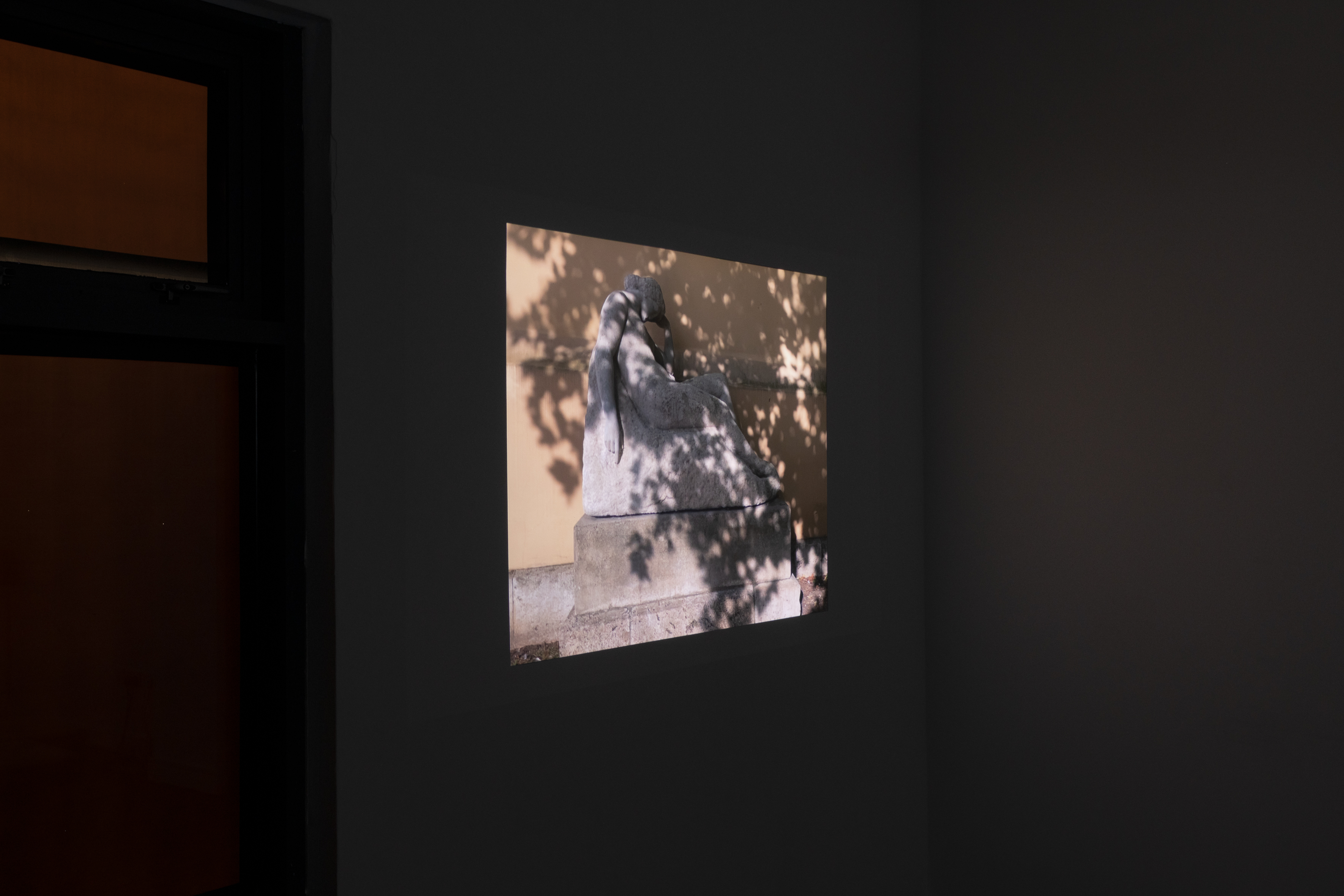 A small projected film still on a dark gallery wall showing a reclining stone statue dappled with tree shadow, set beside an open doorway with warm light beyond.
