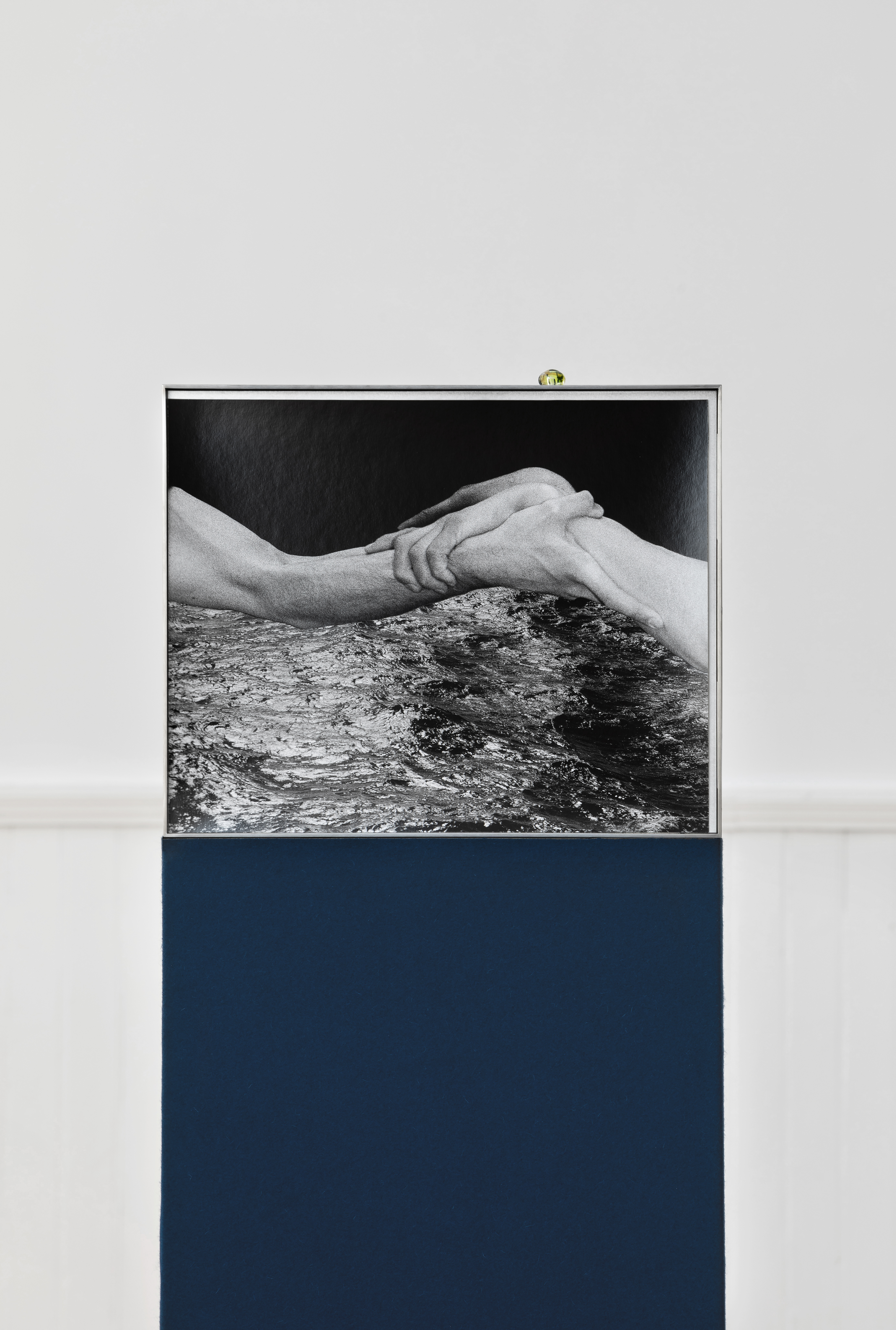 A black and white photograph of two clasped hands held above choppy water, framed in metal and displayed on a navy plinth.