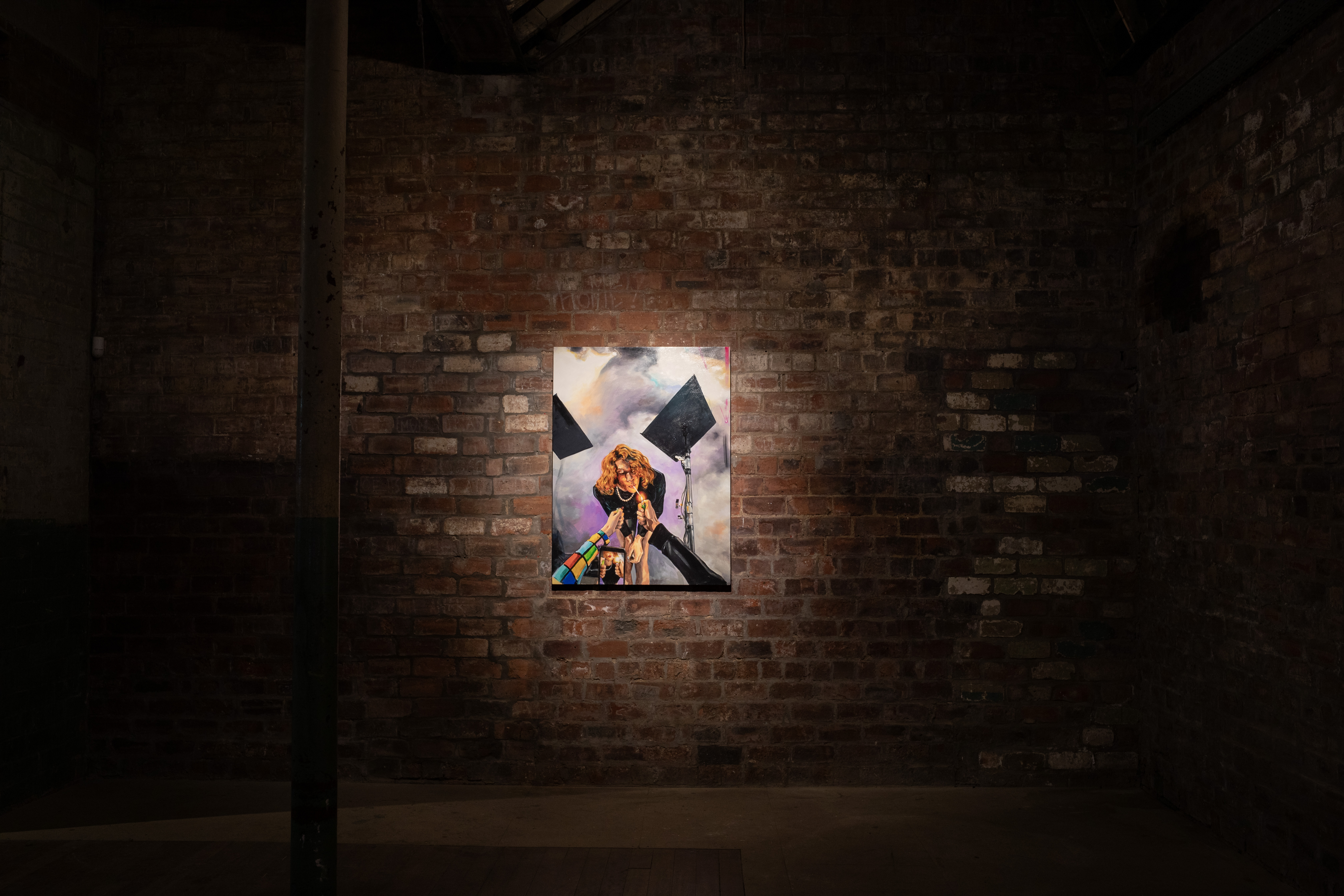 A spotlight illuminates a colourful portrait painting mounted on a weathered brick wall inside a dimly lit industrial gallery space, with exposed brick and a support column framing the installation.