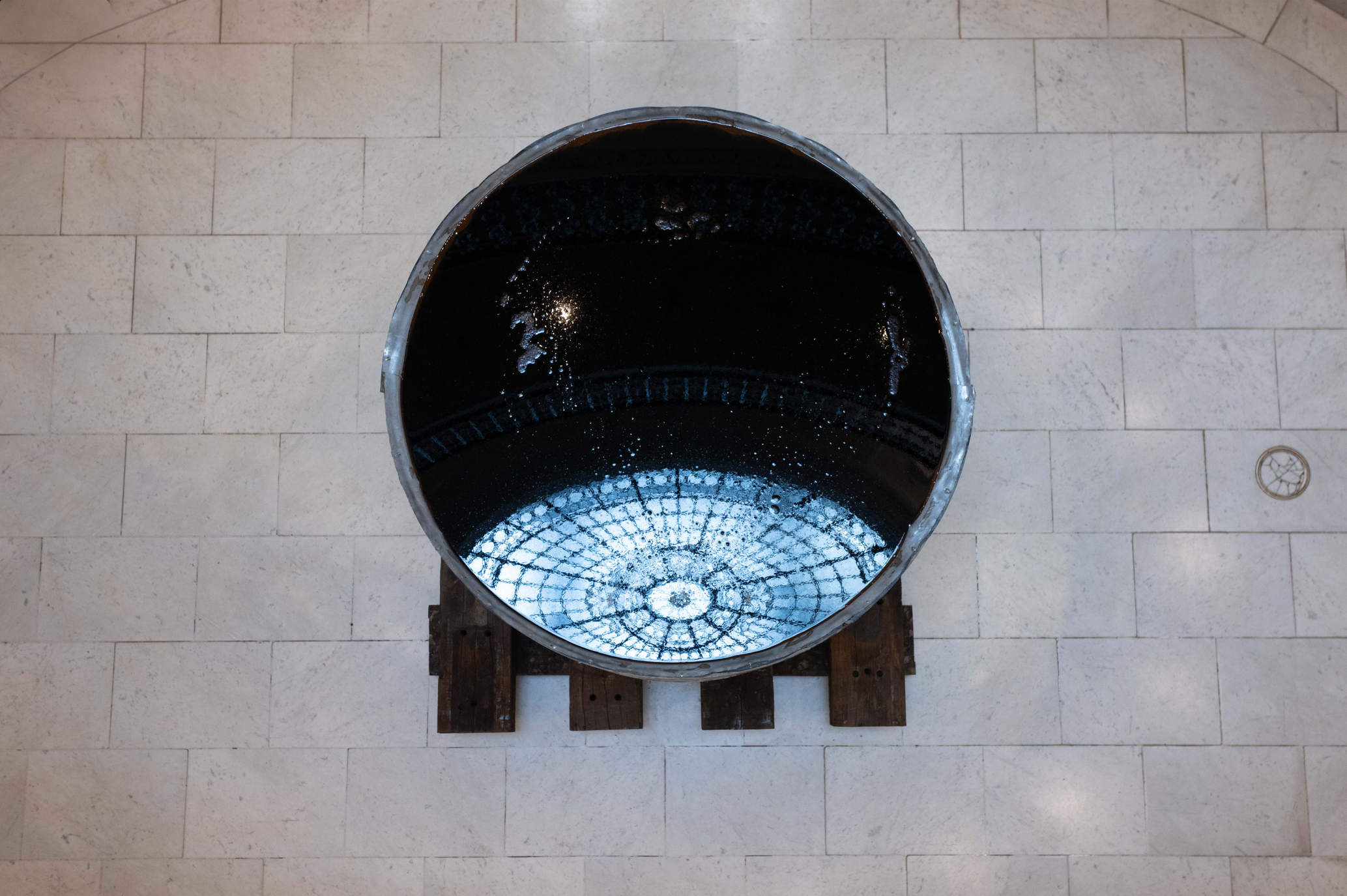 Overhead view of a large circular metal vessel with a dark reflective interior surface on a marble tiled floor, reflecting the domed skylight above it.