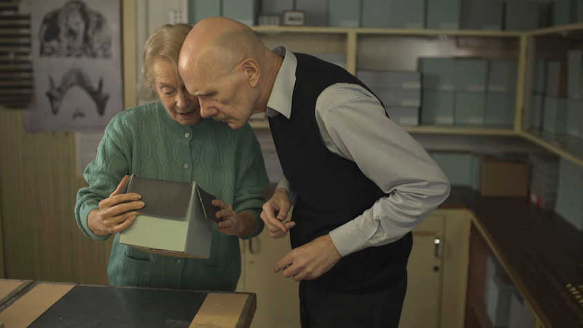 Film still showing two older people looking carefully into a cardboard box. They appear to be in an archive